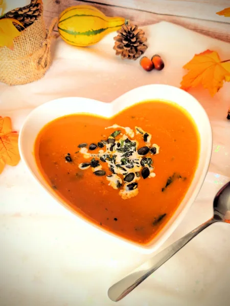 Rezepte Asiatisch inspirierte Kürbissuppe mit Möhren, Süßkartoffel und Kokosmilch in einer herzförmigen weißen Schüssel, garniert mit Kürbiskernen und frischen Kräutern. Im Hintergrund herbstliche Deko mit Kürbis, Tannenzapfen und bunten Blättern.