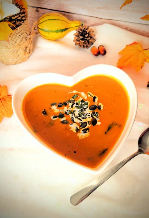 Asiatisch inspirierte Kürbissuppe mit Möhren, Süßkartoffel und Kokosmilch in einer herzförmigen weißen Schüssel, garniert mit Kürbiskernen und frischen Kräutern. Im Hintergrund herbstliche Deko mit Kürbis, Tannenzapfen und bunten Blättern.