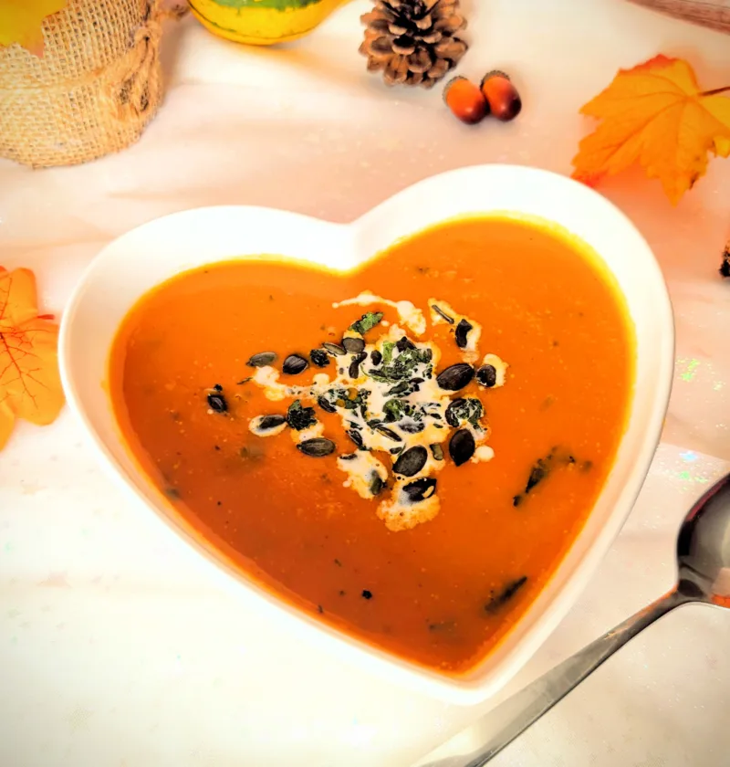 Asiatisch inspirierte Kürbissuppe mit Möhren, Süßkartoffel und Kokosmilch in einer herzförmigen weißen Schüssel, garniert mit Kürbiskernen und frischen Kräutern. Im Hintergrund herbstliche Deko mit Kürbis, Tannenzapfen und bunten Blättern.