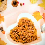 Herbstliches Kürbiskuchen-Porridge in einer weißen herzförmigen Schale, getoppt mit Pekannüssen und Zimt, umgeben von herbstlicher Deko wie bunten Blättern, kleinen Kürbissen und Tannenzapfen; warmes, gemütliches Frühstücksrezept für die Lutealphase.
