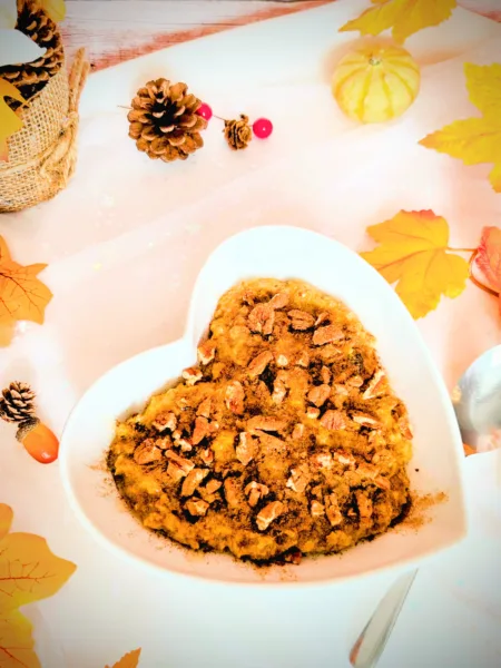 Herbstliches Kürbiskuchen-Porridge in einer weißen herzförmigen Schale, getoppt mit Pekannüssen und Zimt, umgeben von herbstlicher Deko wie bunten Blättern, kleinen Kürbissen und Tannenzapfen; warmes, gemütliches Frühstücksrezept für die Lutealphase.