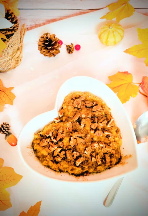 Herbstliches Kürbiskuchen-Porridge in einer weißen herzförmigen Schale, getoppt mit Pekannüssen und Zimt, umgeben von herbstlicher Deko wie bunten Blättern, kleinen Kürbissen und Tannenzapfen; warmes, gemütliches Frühstücksrezept für die Lutealphase.