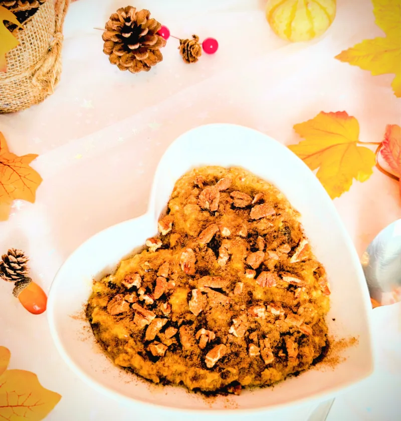 Herbstliches Kürbiskuchen-Porridge in einer weißen herzförmigen Schale, getoppt mit Pekannüssen und Zimt, umgeben von herbstlicher Deko wie bunten Blättern, kleinen Kürbissen und Tannenzapfen; warmes, gemütliches Frühstücksrezept für die Lutealphase.