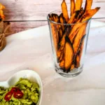 Knusprige Süßkartoffel-Pommes im Glas, serviert mit cremiger Guacamole und frischen Chilischeiben. Die goldbraun gebackenen Süßkartoffel-Pommes wurden im Ofen zubereitet und stehen auf einer hellen Tischdecke vor einer rustikalen Holzrückwand.
