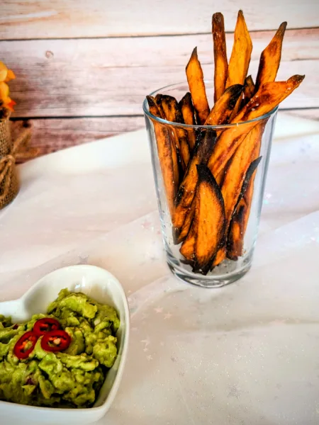 Knusprige Süßkartoffel-Pommes im Glas, serviert mit cremiger Guacamole und frischen Chilischeiben. Die goldbraun gebackenen Süßkartoffel-Pommes wurden im Ofen zubereitet und stehen auf einer hellen Tischdecke vor einer rustikalen Holzrückwand.