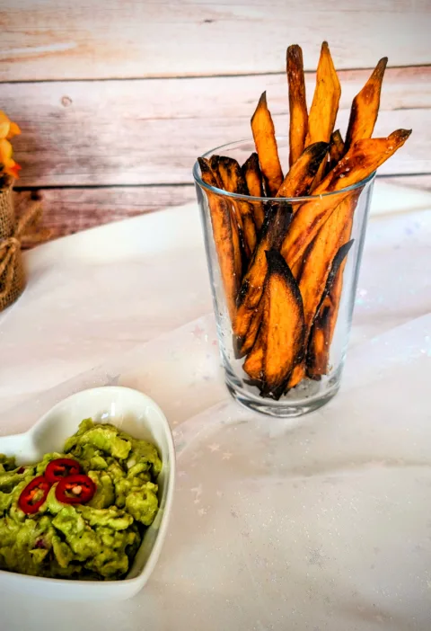 Knusprige Süßkartoffel-Pommes im Glas, serviert mit cremiger Guacamole und frischen Chilischeiben. Die goldbraun gebackenen Süßkartoffel-Pommes wurden im Ofen zubereitet und stehen auf einer hellen Tischdecke vor einer rustikalen Holzrückwand.