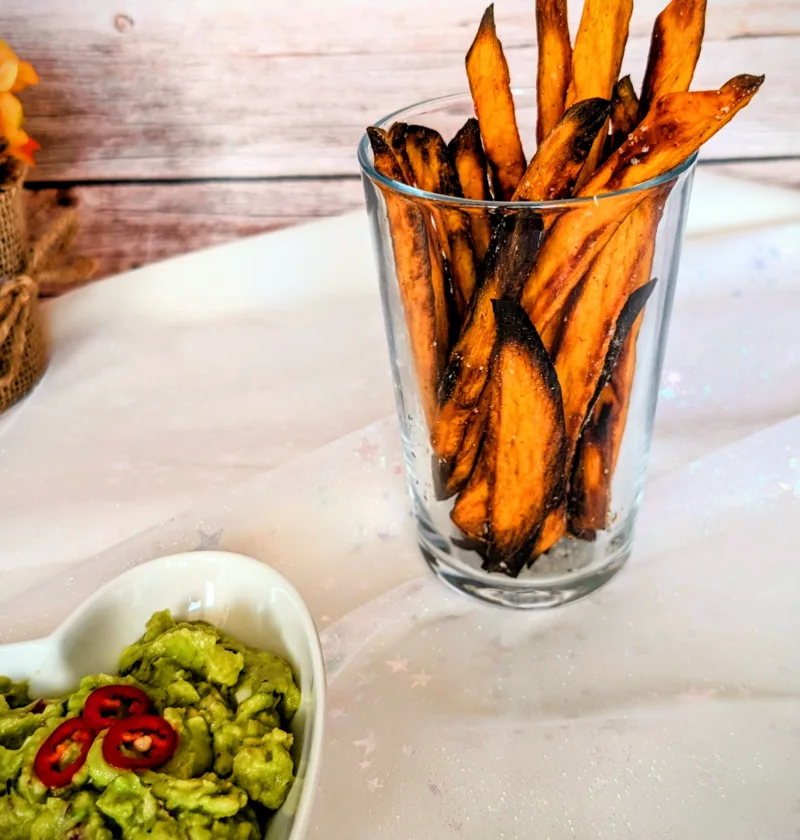 Knusprige Süßkartoffel-Pommes im Glas, serviert mit cremiger Guacamole und frischen Chilischeiben. Die goldbraun gebackenen Süßkartoffel-Pommes wurden im Ofen zubereitet und stehen auf einer hellen Tischdecke vor einer rustikalen Holzrückwand.