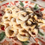 Ein roter Teller voller frisch gebackener Vanillekipferl, bestäubt mit Vanillepuderzucker, arrangiert auf weihnachtlich bedruckten Servietten. Im Hintergrund stehen kleine dekorative Tannenbäume, Christbaumschmuck und weitere festliche Elemente, die eine gemütliche Adventsstimmung vermitteln.