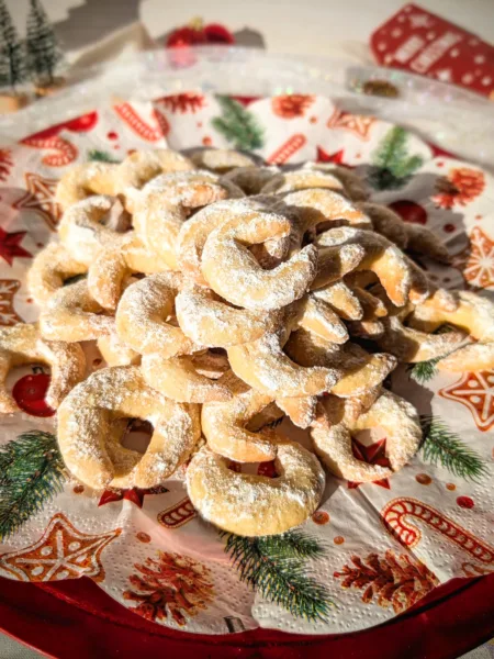 Ein roter Teller voller frisch gebackener Vanillekipferl, bestäubt mit Vanillepuderzucker, arrangiert auf weihnachtlich bedruckten Servietten. Im Hintergrund stehen kleine dekorative Tannenbäume, Christbaumschmuck und weitere festliche Elemente, die eine gemütliche Adventsstimmung vermitteln.