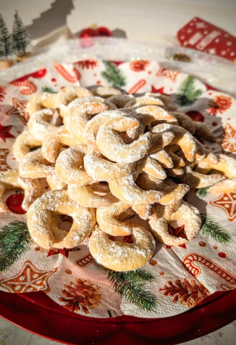 Ein roter Teller voller frisch gebackener Vanillekipferl, bestäubt mit Vanillepuderzucker, arrangiert auf weihnachtlich bedruckten Servietten. Im Hintergrund stehen kleine dekorative Tannenbäume, Christbaumschmuck und weitere festliche Elemente, die eine gemütliche Adventsstimmung vermitteln.