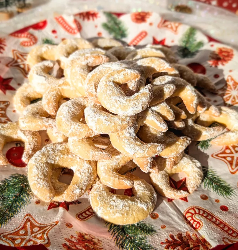 Ein roter Teller voller frisch gebackener Vanillekipferl, bestäubt mit Vanillepuderzucker, arrangiert auf weihnachtlich bedruckten Servietten. Im Hintergrund stehen kleine dekorative Tannenbäume, Christbaumschmuck und weitere festliche Elemente, die eine gemütliche Adventsstimmung vermitteln.