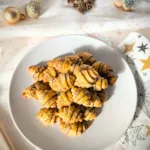 Goldbraun gebackene Zimthörnchen-Plätzchen in Croissant-Form auf einem weißen Teller, dekoriert mit weihnachtlichen Elementen wie Tannenzapfen, kleinen Tannenbäumen und Christbaumkugeln auf hellem Stoffhintergrund.