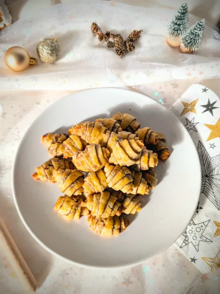 Goldbraun gebackene Zimthörnchen-Plätzchen in Croissant-Form auf einem weißen Teller, dekoriert mit weihnachtlichen Elementen wie Tannenzapfen, kleinen Tannenbäumen und Christbaumkugeln auf hellem Stoffhintergrund.