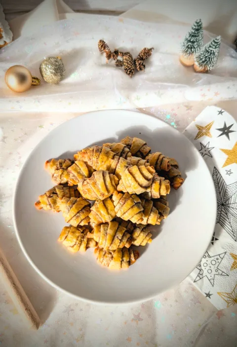Goldbraun gebackene Zimthörnchen-Plätzchen in Croissant-Form auf einem weißen Teller, dekoriert mit weihnachtlichen Elementen wie Tannenzapfen, kleinen Tannenbäumen und Christbaumkugeln auf hellem Stoffhintergrund.