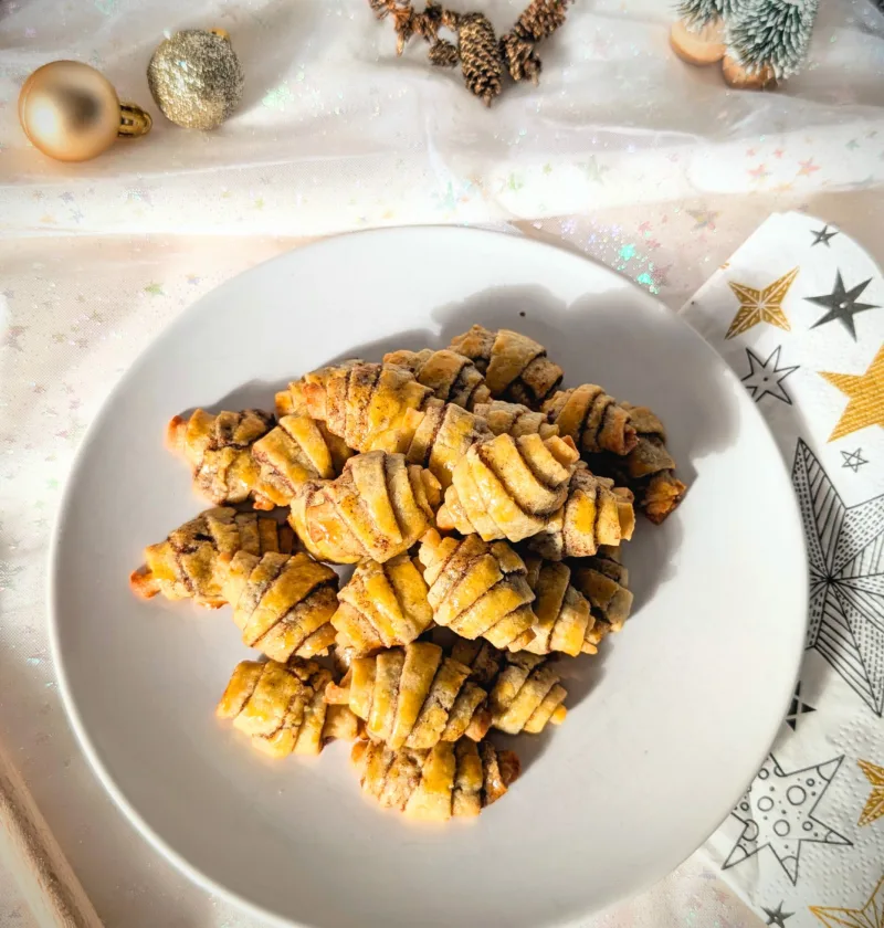 Goldbraun gebackene Zimthörnchen-Plätzchen in Croissant-Form auf einem weißen Teller, dekoriert mit weihnachtlichen Elementen wie Tannenzapfen, kleinen Tannenbäumen und Christbaumkugeln auf hellem Stoffhintergrund.