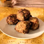 Saftige Hackbällchen aus gemischtem Hackfleisch auf einem weißen Teller, goldbraun gebraten und locker aufgeschichtet, fotografiert auf gelbem Stoff vor hellem Holz-Hintergrund.