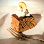 Saftige Protein-Zimt-Blondies aus weißen Bohnen, auf einem weißen Teller serviert, getoppt mit einem Klecks Skyr und einer Bananenscheibe, fotografiert bei warmem Tageslicht.