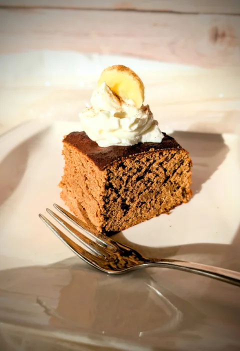 Saftige Protein-Zimt-Blondies aus weißen Bohnen, auf einem weißen Teller serviert, getoppt mit einem Klecks Skyr und einer Bananenscheibe, fotografiert bei warmem Tageslicht.