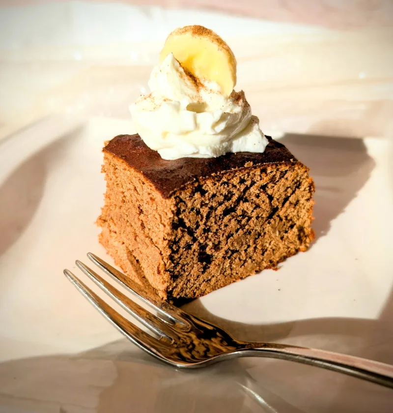 Saftige Protein-Zimt-Blondies aus weißen Bohnen, auf einem weißen Teller serviert, getoppt mit einem Klecks Skyr und einer Bananenscheibe, fotografiert bei warmem Tageslicht.