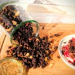 Hausgemachtes Schokoladen-Granola aus Haferflocken, Nüssen und Samen, aus einem Glas auf ein Holzbrett geschüttet und mit Joghurt und Beeren serviert.