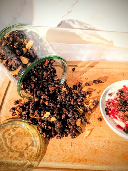 Hausgemachtes Schokoladen-Granola aus Haferflocken, Nüssen und Samen, aus einem Glas auf ein Holzbrett geschüttet und mit Joghurt und Beeren serviert.