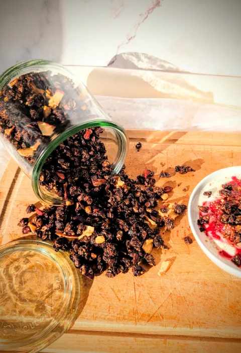 Hausgemachtes Schokoladen-Granola aus Haferflocken, Nüssen und Samen, aus einem Glas auf ein Holzbrett geschüttet und mit Joghurt und Beeren serviert.