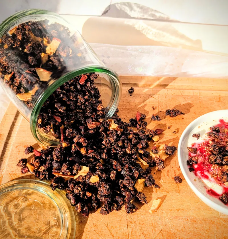 Hausgemachtes Schokoladen-Granola aus Haferflocken, Nüssen und Samen, aus einem Glas auf ein Holzbrett geschüttet und mit Joghurt und Beeren serviert.