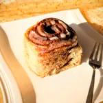 Sauerteig-Zimtschnecke auf weißem Teller, spiralförmige Zimtfüllung von oben sichtbar, mit Zuckerguss und weicher Teigstruktur, fotografiert im warmen Licht.