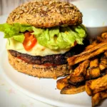 Selbstgemachter Smash-Cheeseburger mit reinem Rindfleisch-Patty im Vollkorn-Körner-Bun, belegt mit geschmolzenem Käse, frischem Salat und Tomaten, serviert mit knusprigen Süßkartoffelpommes auf weißem Teller.