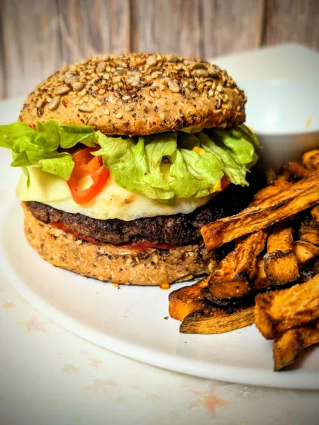Selbstgemachter Smash-Cheeseburger mit reinem Rindfleisch-Patty im Vollkorn-Körner-Bun, belegt mit geschmolzenem Käse, frischem Salat und Tomaten, serviert mit knusprigen Süßkartoffelpommes auf weißem Teller.