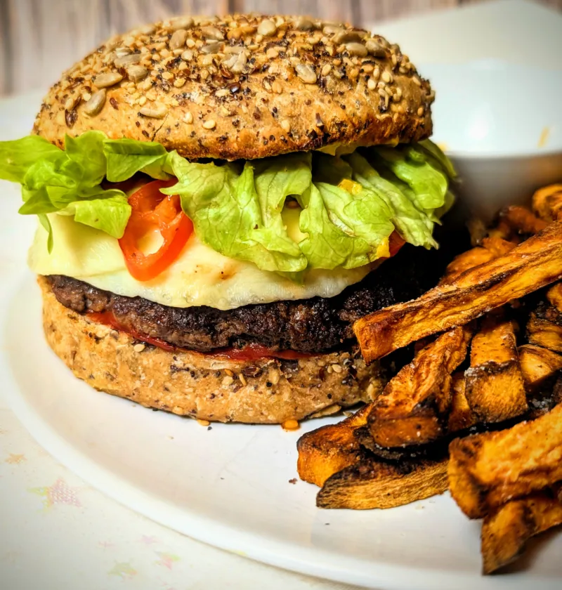 Selbstgemachter Smash-Cheeseburger mit reinem Rindfleisch-Patty im Vollkorn-Körner-Bun, belegt mit geschmolzenem Käse, frischem Salat und Tomaten, serviert mit knusprigen Süßkartoffelpommes auf weißem Teller.