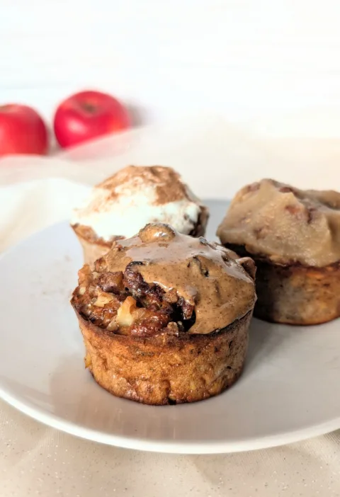 Saftige Apfel-Frühstücksmuffins mit Haferflocken auf einem weißen Teller, getoppt mit Mandelmus, Frischkäse und Apfelmark, frische Äpfel im Hintergrund – gesundes Frühstücksrezept für den Herbst.