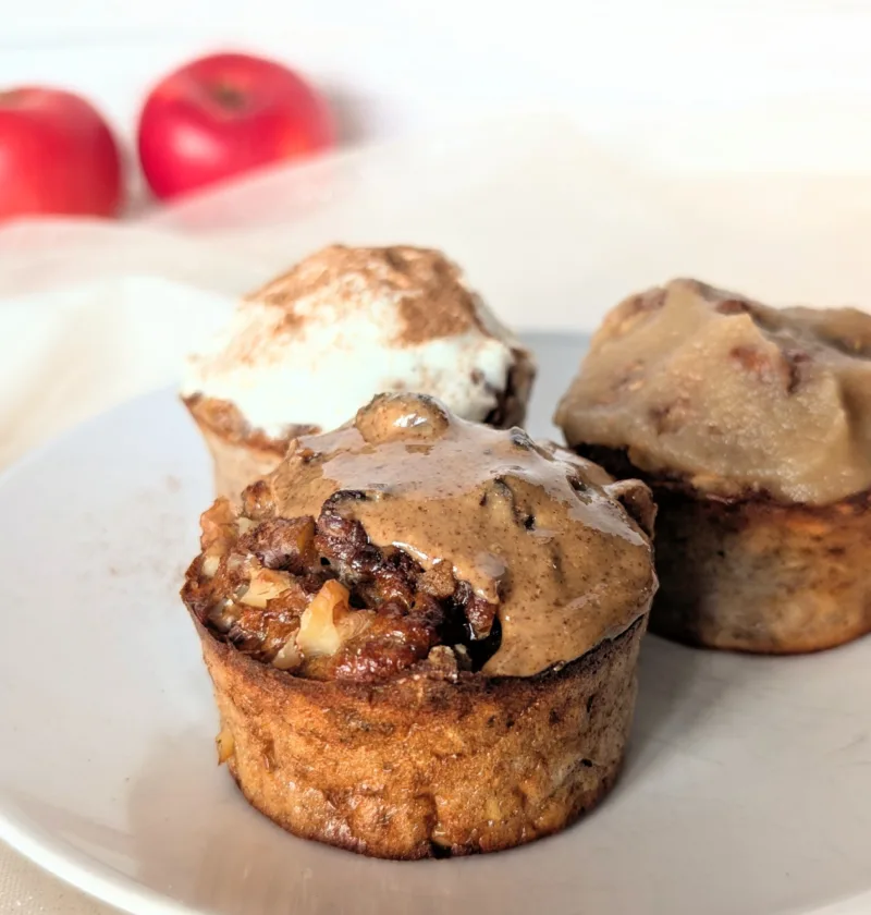 Saftige Apfel-Frühstücksmuffins mit Haferflocken auf einem weißen Teller, getoppt mit Mandelmus, Frischkäse und Apfelmark, frische Äpfel im Hintergrund – gesundes Frühstücksrezept für den Herbst.