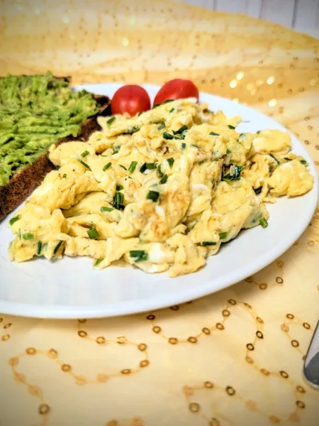 Teller mit lockerem Rührei mit frischem Schnittlauch, serviert mit einer Scheibe Sauerteig-Vollkornbrot mit Avocado und zwei Cherrytomaten – ein einfaches, proteinreiches Frühstück in natürlichem Licht fotografiert.