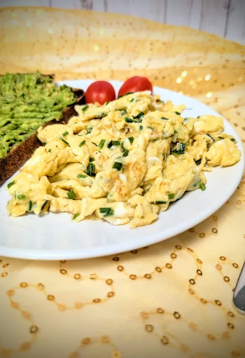 Teller mit lockerem Rührei mit frischem Schnittlauch, serviert mit einer Scheibe Sauerteig-Vollkornbrot mit Avocado und zwei Cherrytomaten – ein einfaches, proteinreiches Frühstück in natürlichem Licht fotografiert.
