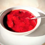 Selbstgemachtes Erdbeerkompott mit ganzen Erdbeerstücken in einer weißen Schale, serviert auf einem Teller im warmen Sonnenlicht.