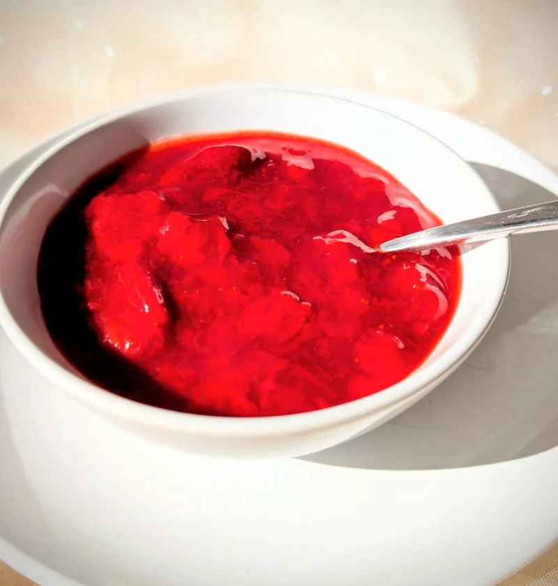 Selbstgemachtes Erdbeerkompott mit ganzen Erdbeerstücken in einer weißen Schale, serviert auf einem Teller im warmen Sonnenlicht.