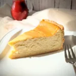 Ein Stück klassischer Käsekuchen mit goldgelber Oberfläche und Mürbeteigboden auf einem weißen Teller, serviert mit einer Kanne roter Erdbeersauce im Hintergrund.