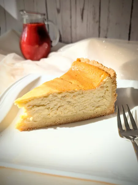 Ein Stück klassischer Käsekuchen mit goldgelber Oberfläche und Mürbeteigboden auf einem weißen Teller, serviert mit einer Kanne roter Erdbeersauce im Hintergrund.
