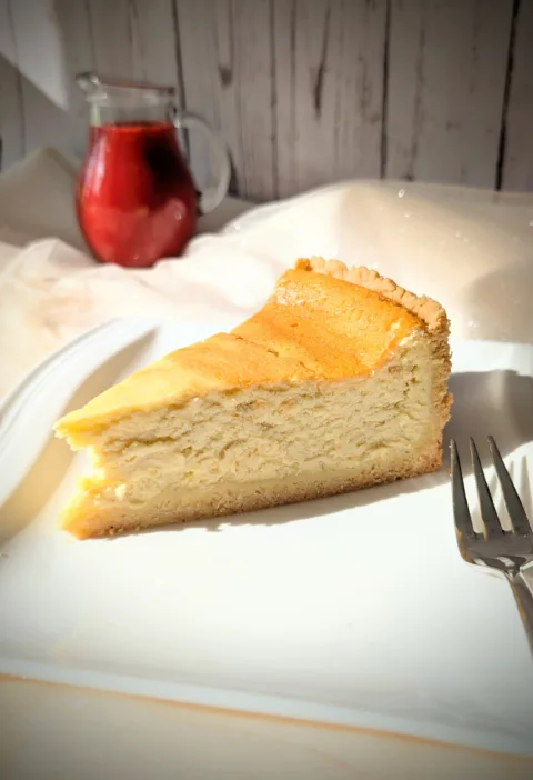 Ein Stück klassischer Käsekuchen mit goldgelber Oberfläche und Mürbeteigboden auf einem weißen Teller, serviert mit einer Kanne roter Erdbeersauce im Hintergrund.