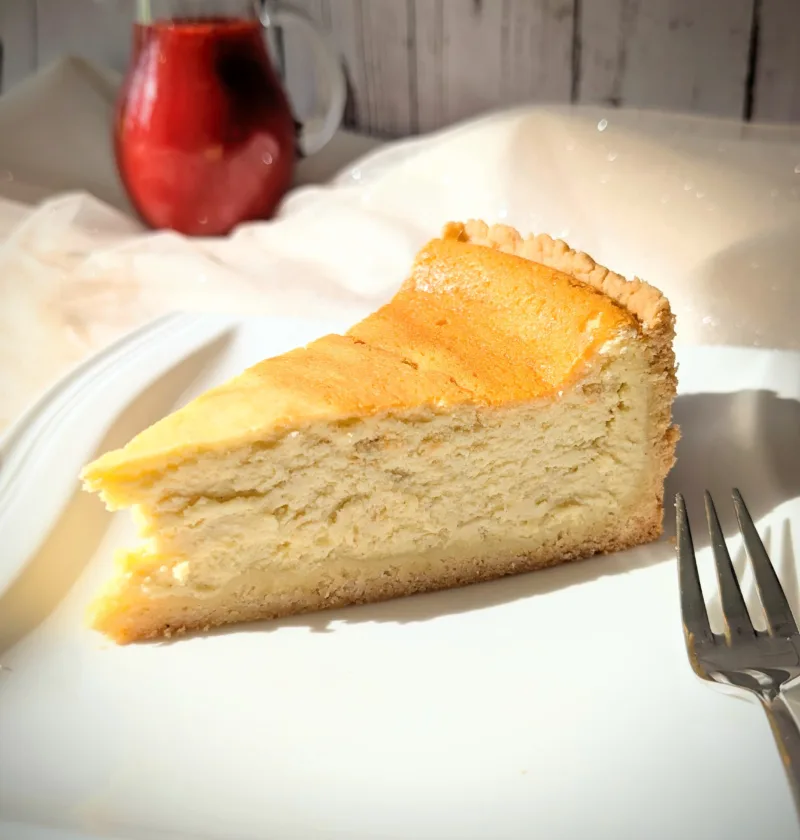 Ein Stück klassischer Käsekuchen mit goldgelber Oberfläche und Mürbeteigboden auf einem weißen Teller, serviert mit einer Kanne roter Erdbeersauce im Hintergrund.