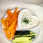 Cremige Sour Cream Alternative aus Frischkäse und Magerquark in einer kleinen Schale, mit Schnittlauch und Pfeffer verfeinert, serviert mit frischen Paprikastreifen und Salatgurke auf einem rustikalen Hintergrund.