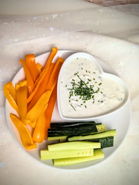 Cremige Sour Cream Alternative aus Frischkäse und Magerquark in einer kleinen Schale, mit Schnittlauch und Pfeffer verfeinert, serviert mit frischen Paprikastreifen und Salatgurke auf einem rustikalen Hintergrund.