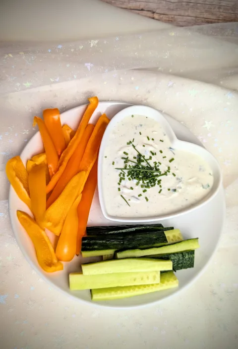 Cremige Sour Cream Alternative aus Frischkäse und Magerquark in einer kleinen Schale, mit Schnittlauch und Pfeffer verfeinert, serviert mit frischen Paprikastreifen und Salatgurke auf einem rustikalen Hintergrund.