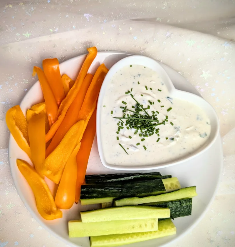 Cremige Sour Cream Alternative aus Frischkäse und Magerquark in einer kleinen Schale, mit Schnittlauch und Pfeffer verfeinert, serviert mit frischen Paprikastreifen und Salatgurke auf einem rustikalen Hintergrund.