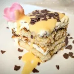 Eierlikör-Tiramisu auf weißem Teller, geschichtet aus Löffelbiskuits und Mascarpone-Creme, mit Eierlikör übergossen und mit Schokoraspeln garniert, dekoriert im frühlingshaften Oster-Setting.