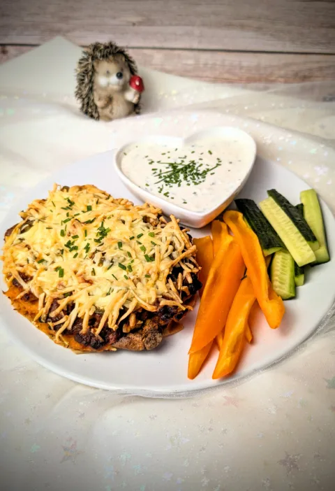Überbackene Süßkartoffel mit würziger BBQ-Hackfleischfüllung, Kichererbsen, Jalapeños und goldbraun geschmolzenem Käse, serviert mit cremigem Dip und frischen Gemüsesticks.