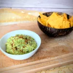 Cremige Guacamole in einer weißen Schale, mit Gewürzen bestreut und auf einem Holzbrett angerichtet. Im Hintergrund steht eine Schale mit knusprigen Tortilla-Chips – ideal zum Dippen und Servieren der selbstgemachten Guacamole.