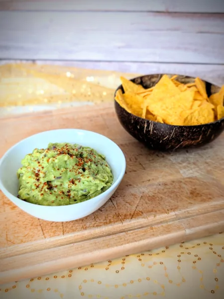 Cremige Guacamole in einer weißen Schale, mit Gewürzen bestreut und auf einem Holzbrett angerichtet. Im Hintergrund steht eine Schale mit knusprigen Tortilla-Chips – ideal zum Dippen und Servieren der selbstgemachten Guacamole.