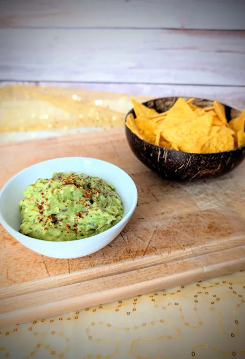 Cremige Guacamole in einer weißen Schale, mit Gewürzen bestreut und auf einem Holzbrett angerichtet. Im Hintergrund steht eine Schale mit knusprigen Tortilla-Chips – ideal zum Dippen und Servieren der selbstgemachten Guacamole.